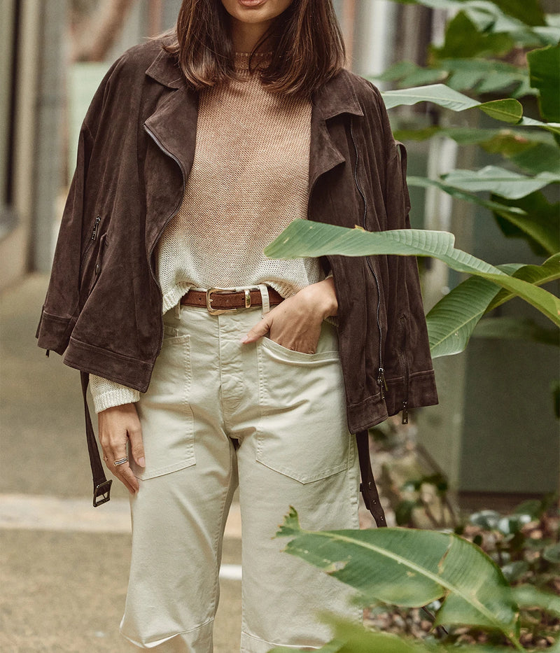 A woman wearing a dark brown suede jacket over a beige knit top and cream cargo pants, styled with a brown belt, standing outdoors in front of green tropical leaves.