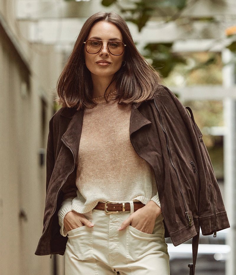A woman wearing round brown sunglasses, a beige knit sweater, cream pants, and a dark brown suede jacket, standing in an outdoor urban setting.