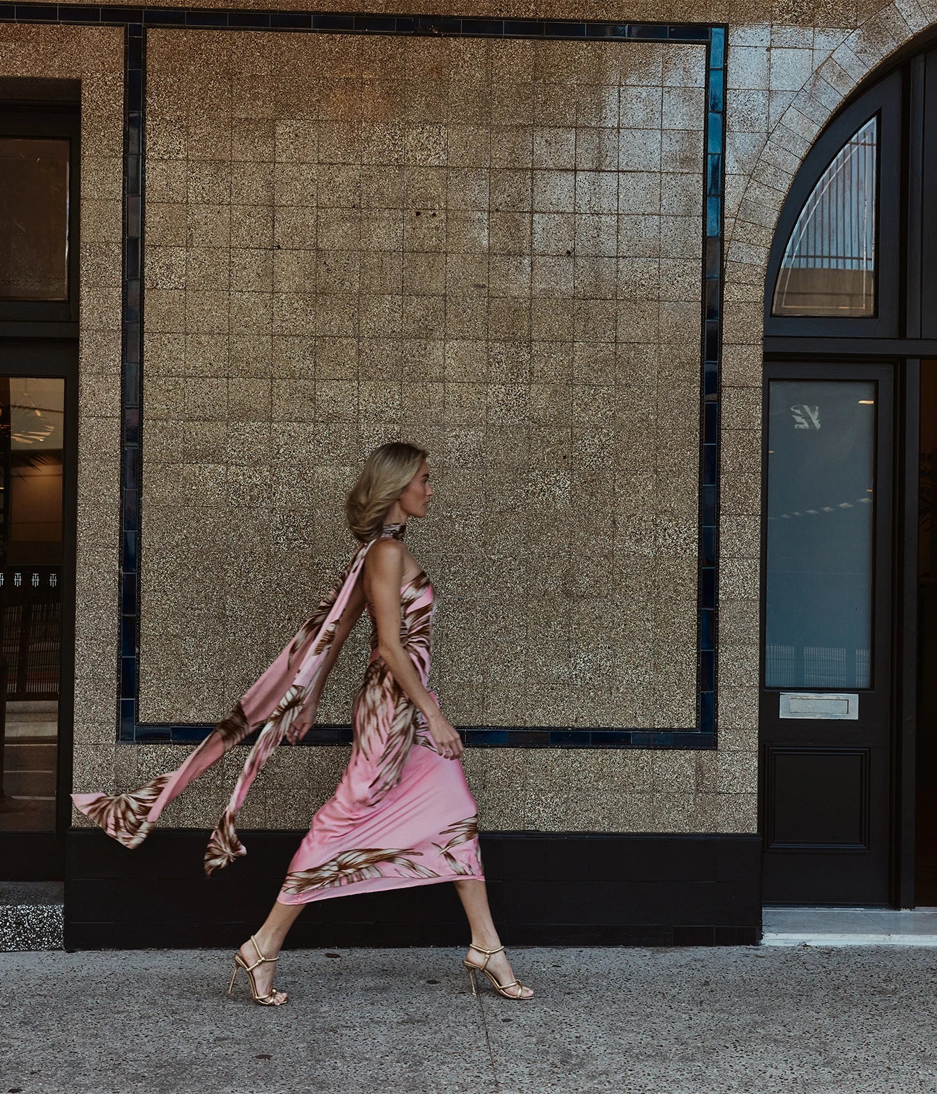 A woman walks in profile wearing a pink sleeveless dress with a metallic gold abstract pattern and a matching sheer scarf, set against a textured beige tiled building facade.