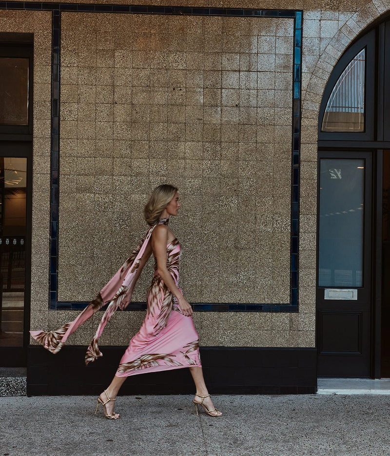 A woman walks in profile wearing a pink sleeveless dress with a metallic gold abstract pattern and a matching sheer scarf, set against a textured beige tiled building facade.