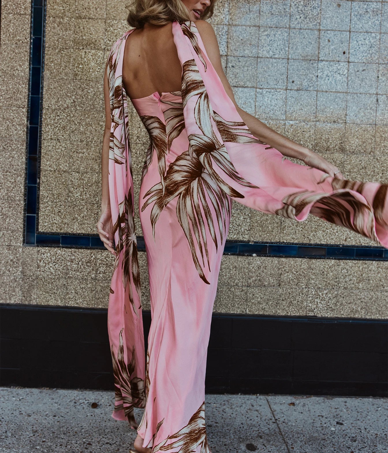 A woman wearing a pink halter-neck maxi dress with a brown palm leaf print, shown from the back and side as she moves, with a tiled wall in the background.