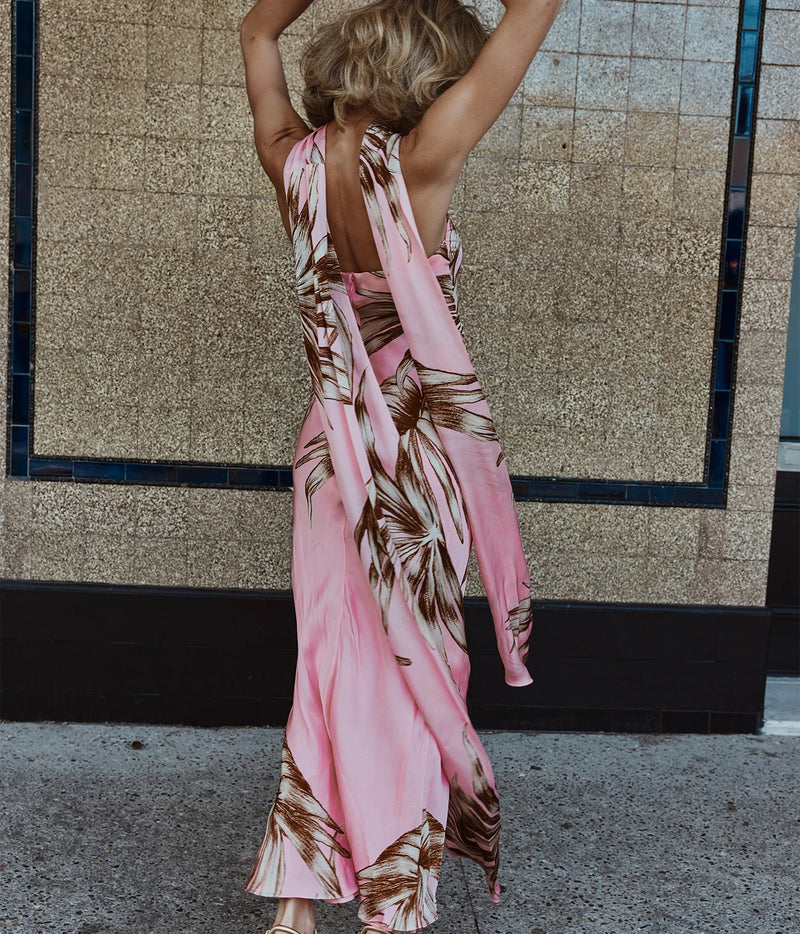 A woman wearing a pink halter maxi dress with a brown palm leaf print, photographed from behind against a tiled wall.