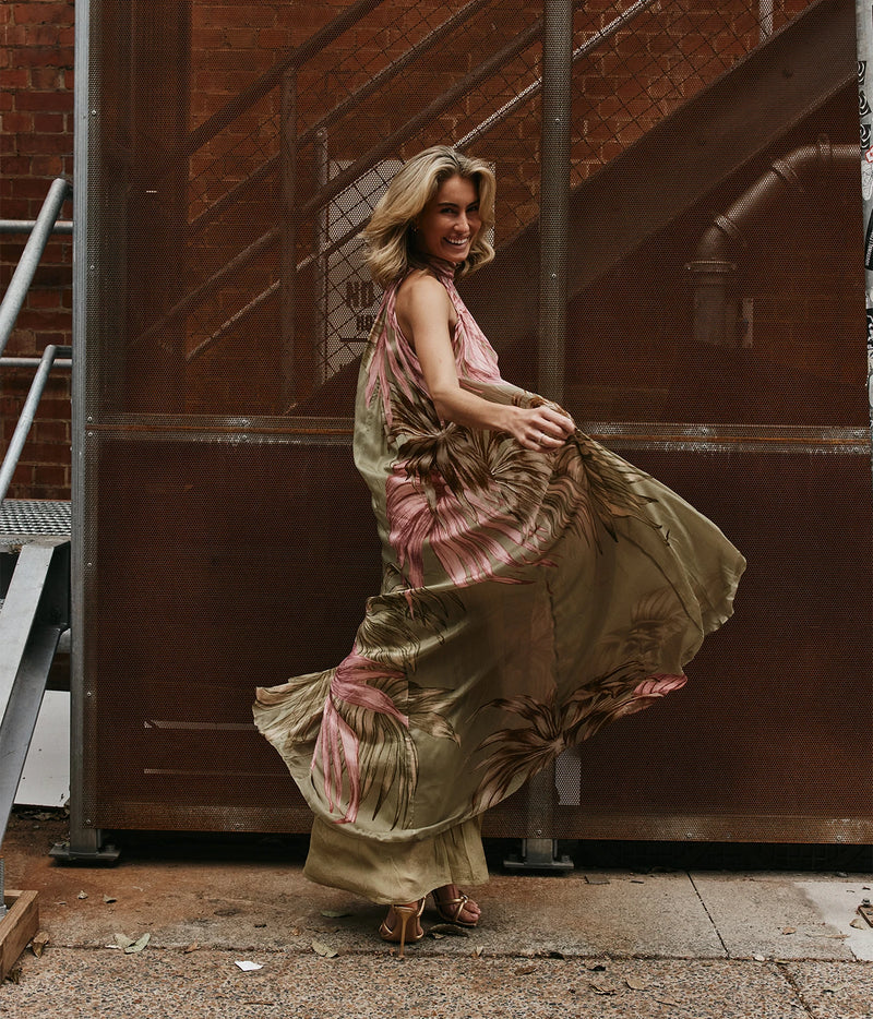 A woman wearing a flowing olive green and pink tropical-print maxi dress and brown strappy heels, captured mid-motion as she twirls in front of a large brown metal gate and brick building.