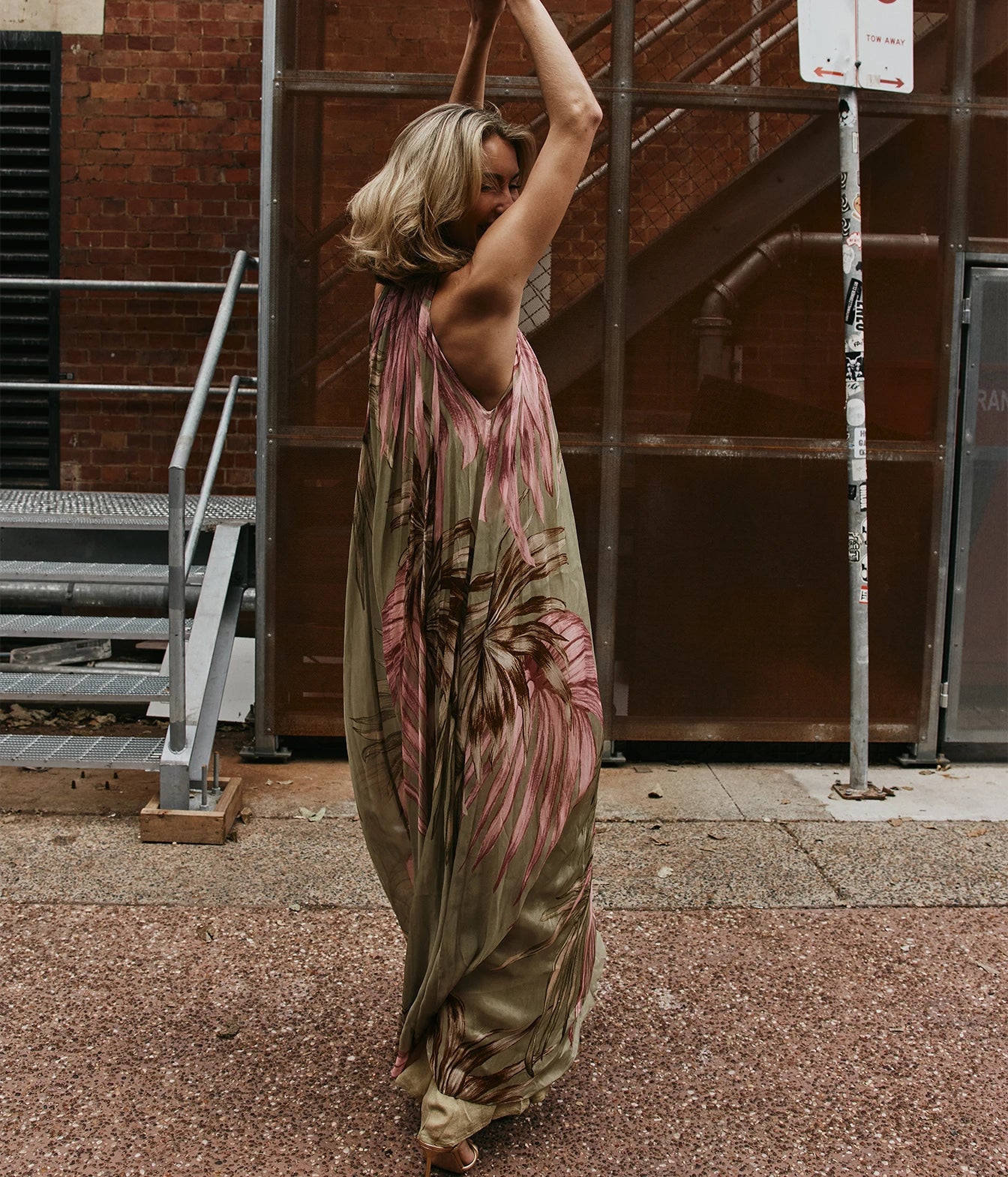 A woman wearing a flowing olive green maxi dress with a pink and brown palm leaf print, standing on a city sidewalk in front of a brick building and metal scaffolding.