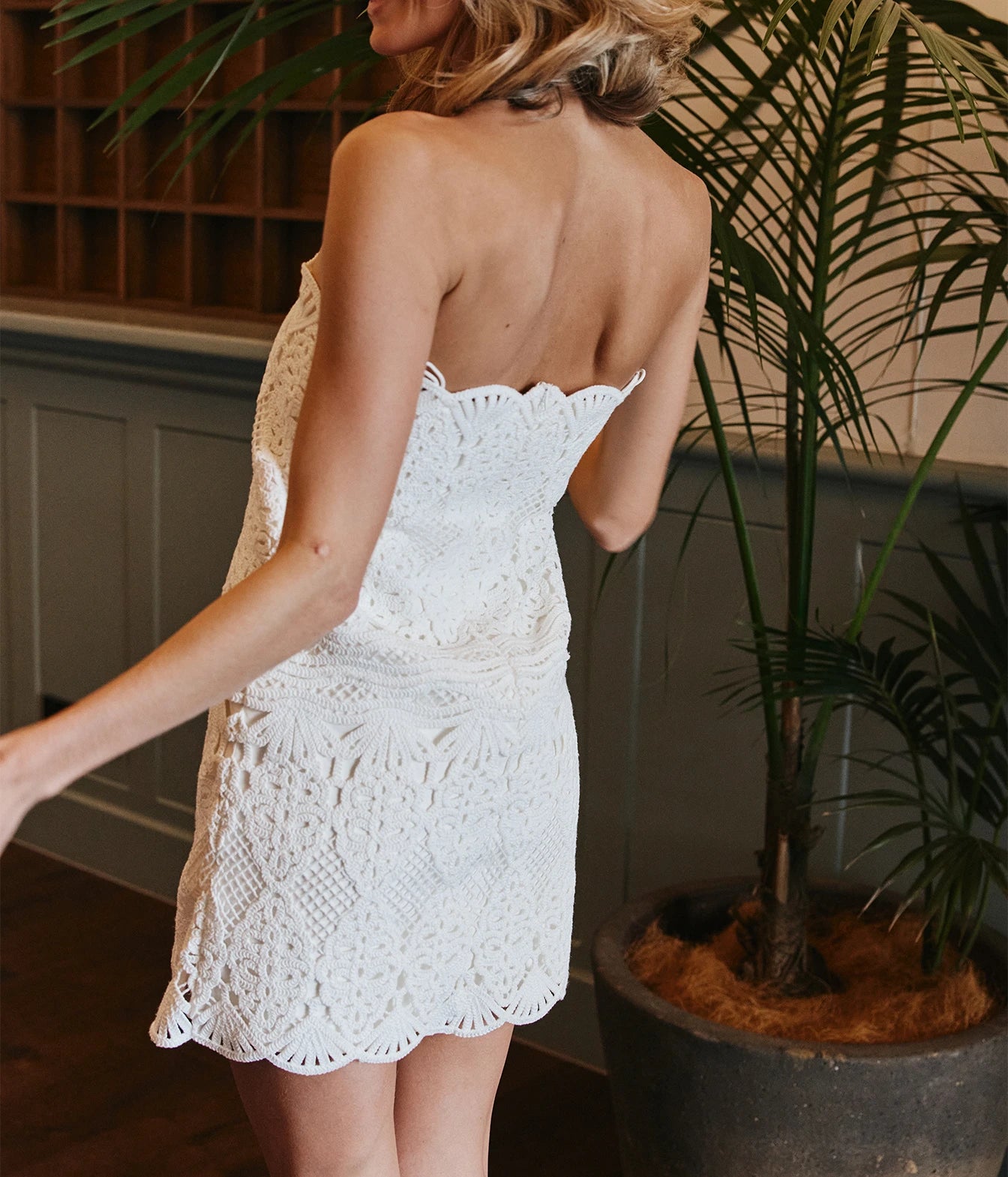 A woman wearing a white crochet strapless mini dress, photographed from behind in an indoor setting with a potted palm plant in the background.
