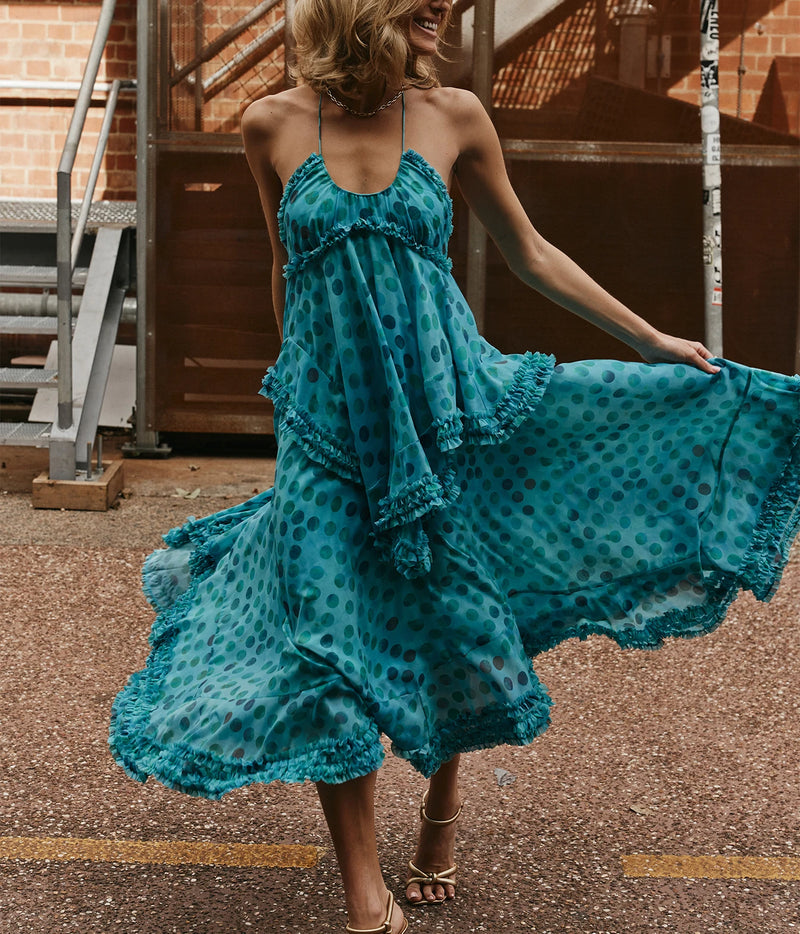 A woman wearing a flowing teal blue polka-dot maxi dress with ruffled layers, captured mid-spin outdoors on a paved street with brick and metal stairs in the background.