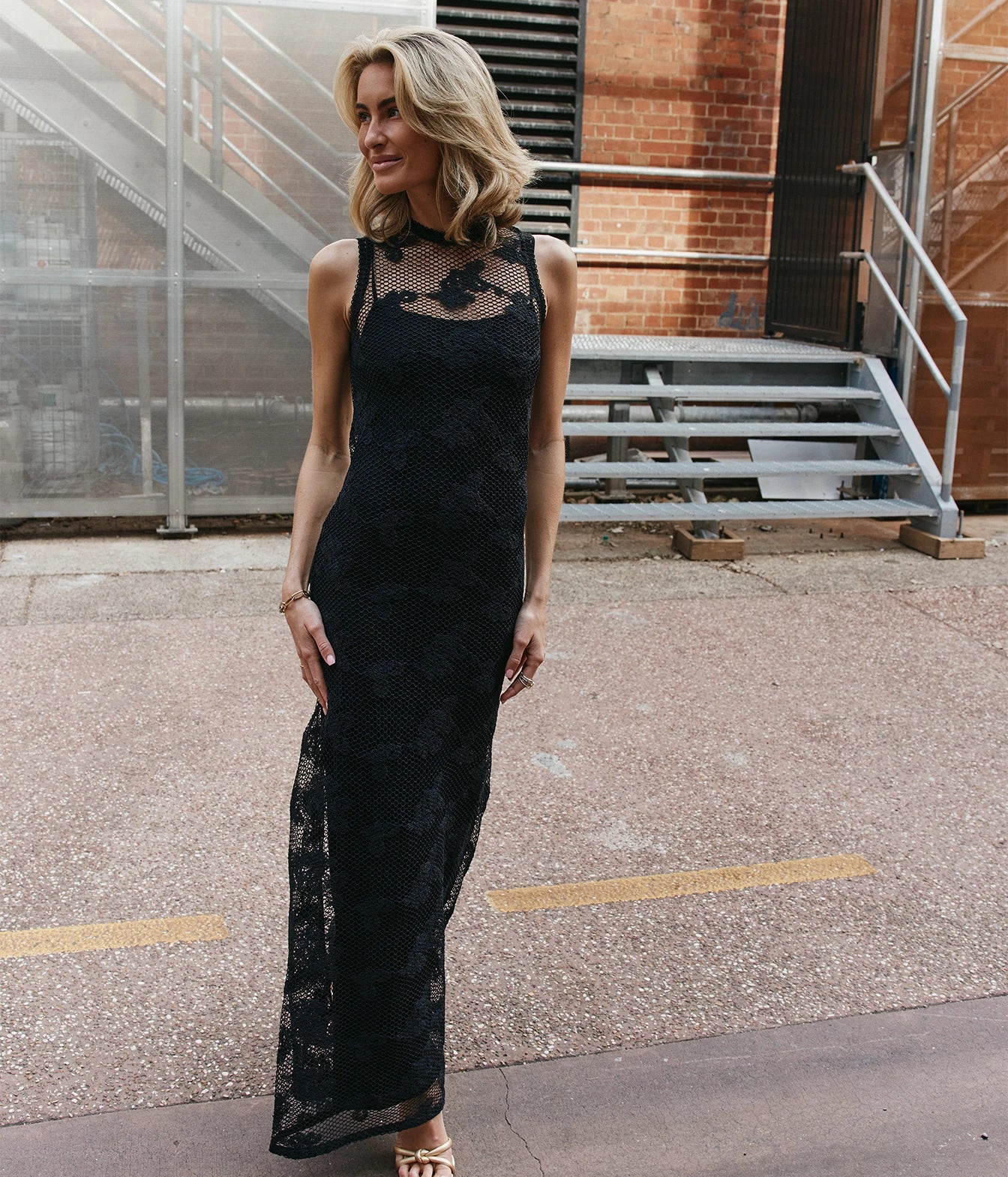 A woman wearing a sleeveless black lace maxi dress with sheer mesh panels at the neckline and upper bodice, standing outdoors on a paved walkway in front of a brick building and metal stairs.