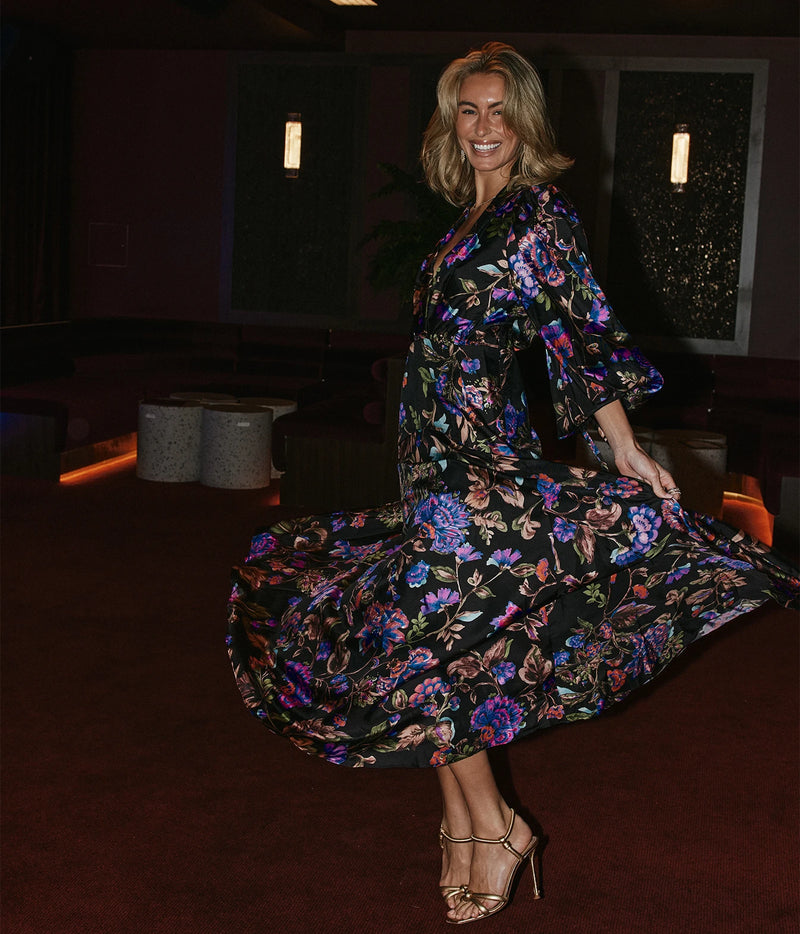 A woman wearing a black midi wrap dress with a vibrant multicolor floral print, twirling in a dimly lit indoor venue.