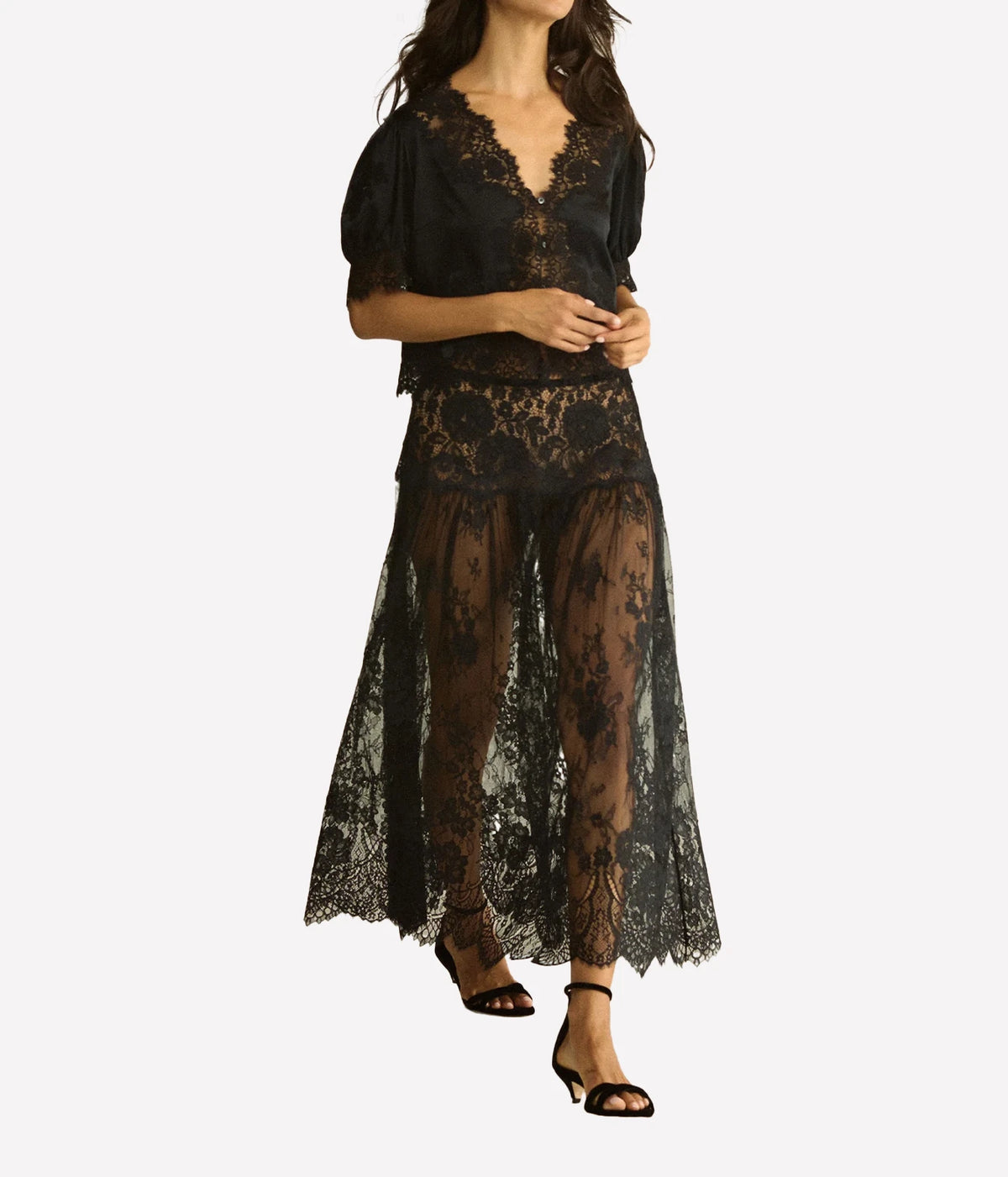 Model walking in the DÔEN Idella Top In Black with a sheer black lace maxi skirt and black ankle-strap sandals, hands clasped at the front