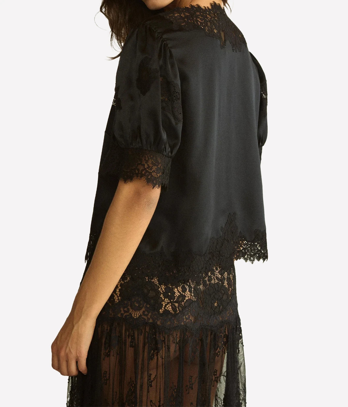 Side view of a model wearing the DÔEN Idella Top In Black with a sheer lace skirt, showing the satin back, puff sleeve and scalloped hem