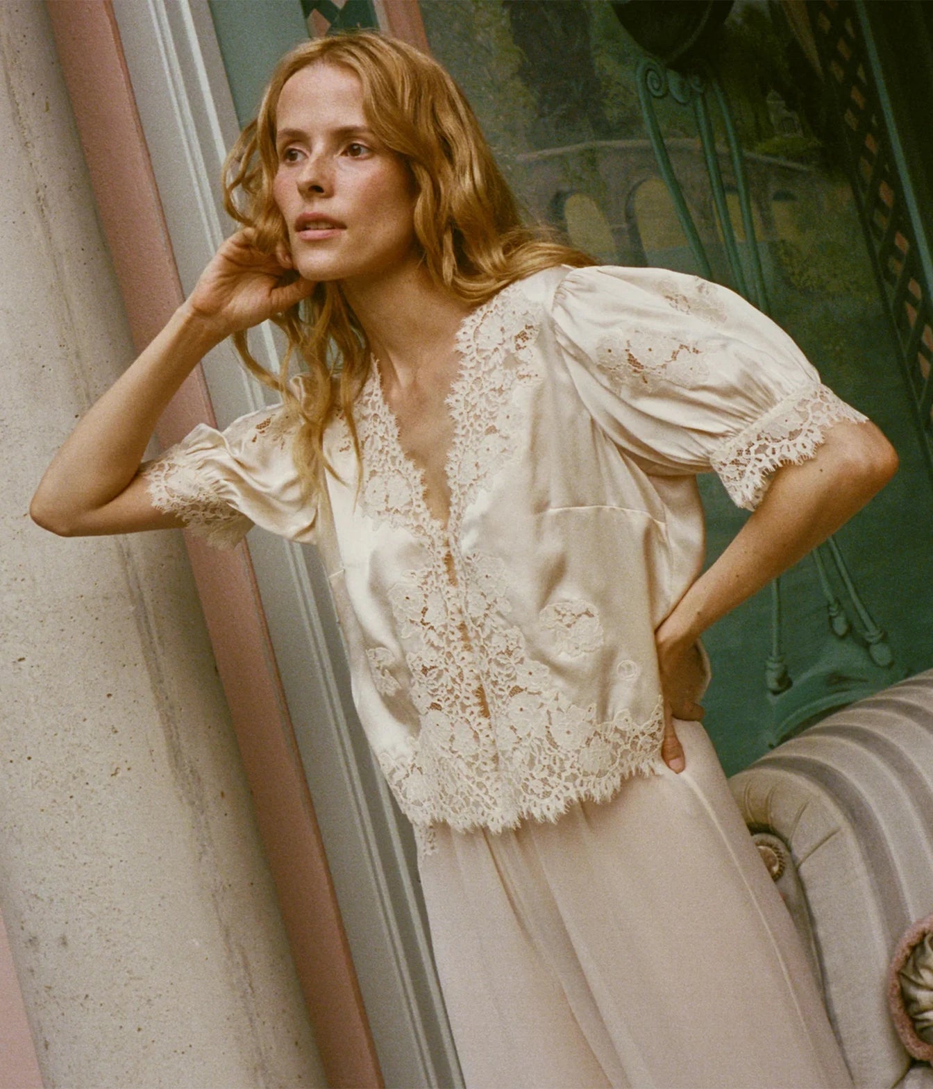 A woman wearing a cream satin short-sleeve blouse with lace trim and a matching cream skirt, posed in front of a rustic interior background.