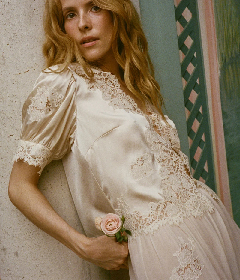 A woman wearing a cream satin slip dress with intricate lace trim and puff sleeves, holding a small pink rose against a textured wall.