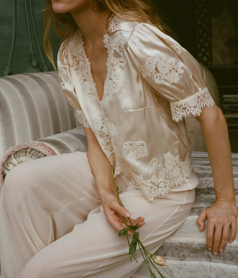 A woman seated on a striped sofa wearing a cream satin pajama set with white lace trim and embroidered floral details. She is holding a small flower stem in a softly lit indoor setting.