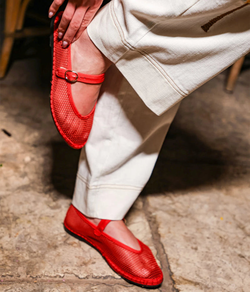 Model wearing the Flabelus Isabella in Red with cream wide-leg trousers, holding one foot up to show the mesh upper and patent leather buckle strap