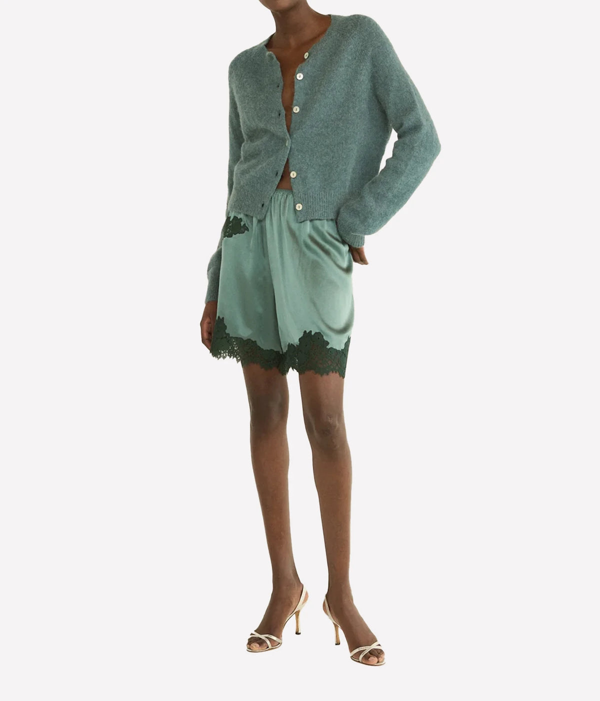 A model wearing a sage green cardigan and matching satin mini skirt with dark green lace trim, paired with beige high-heeled sandals, against a plain light gray background.