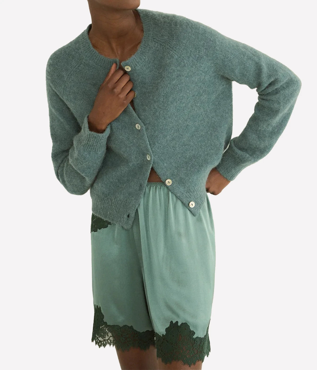 A model wearing a dusty sage green knit cardigan with cream buttons and matching sage green shorts with dark green lace trim, against a plain light gray background.
