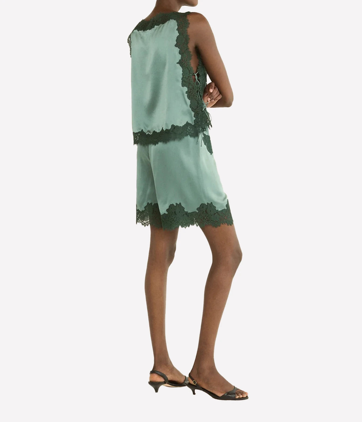 A model shown from the back wearing a sage green sleeveless romper with dark green lace trim, paired with dark brown slingback kitten heels, against a plain light gray background.