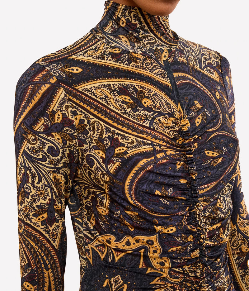 A close-up of a long-sleeve mock-neck top with a ruched front and an intricate paisley pattern in mustard yellow and navy blue, worn by a model.