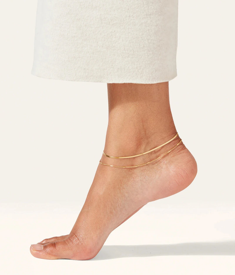 Side view of a model's bare foot raised on her toes wearing the Jenny Bird Surfside Anklet in Gold with cream wide-leg trousers