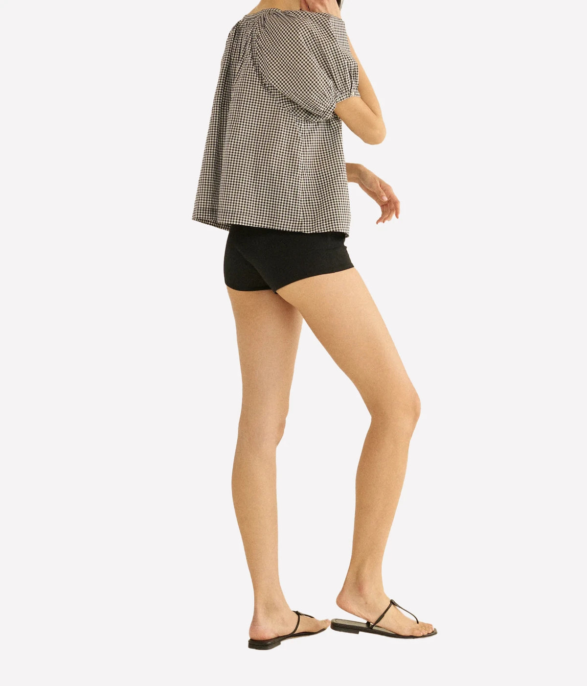 Back view of a model wearing the DÔEN June Top in Noir La Maddalena Gingham with black high-waisted shorts and black thong sandals, showing the full puff sleeves and relaxed back