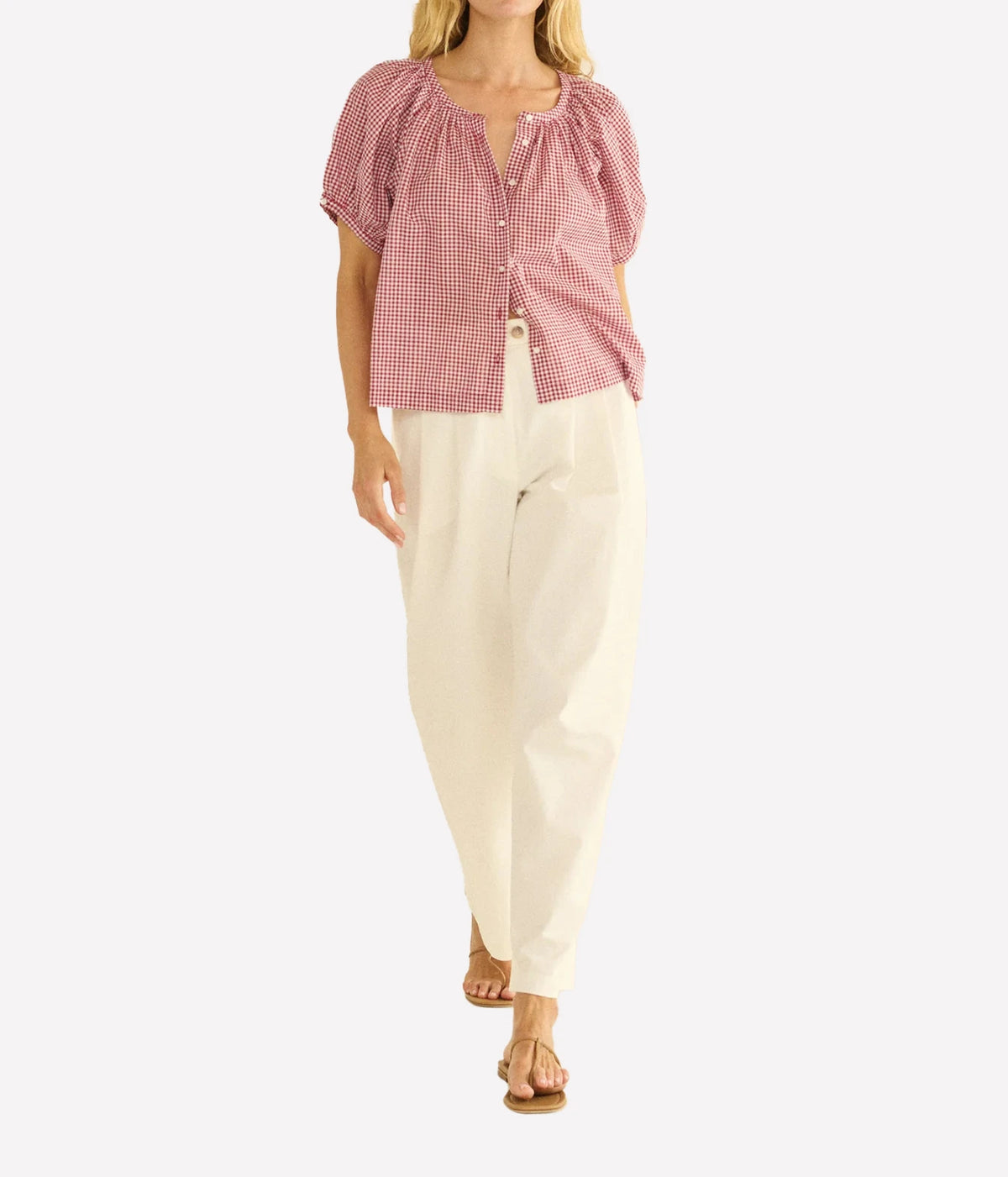 A woman modeling a short-sleeve pink and white gingham blouse with puff sleeves and a split neckline, paired with cream-colored pants and tan sandals, against a plain light gray background.