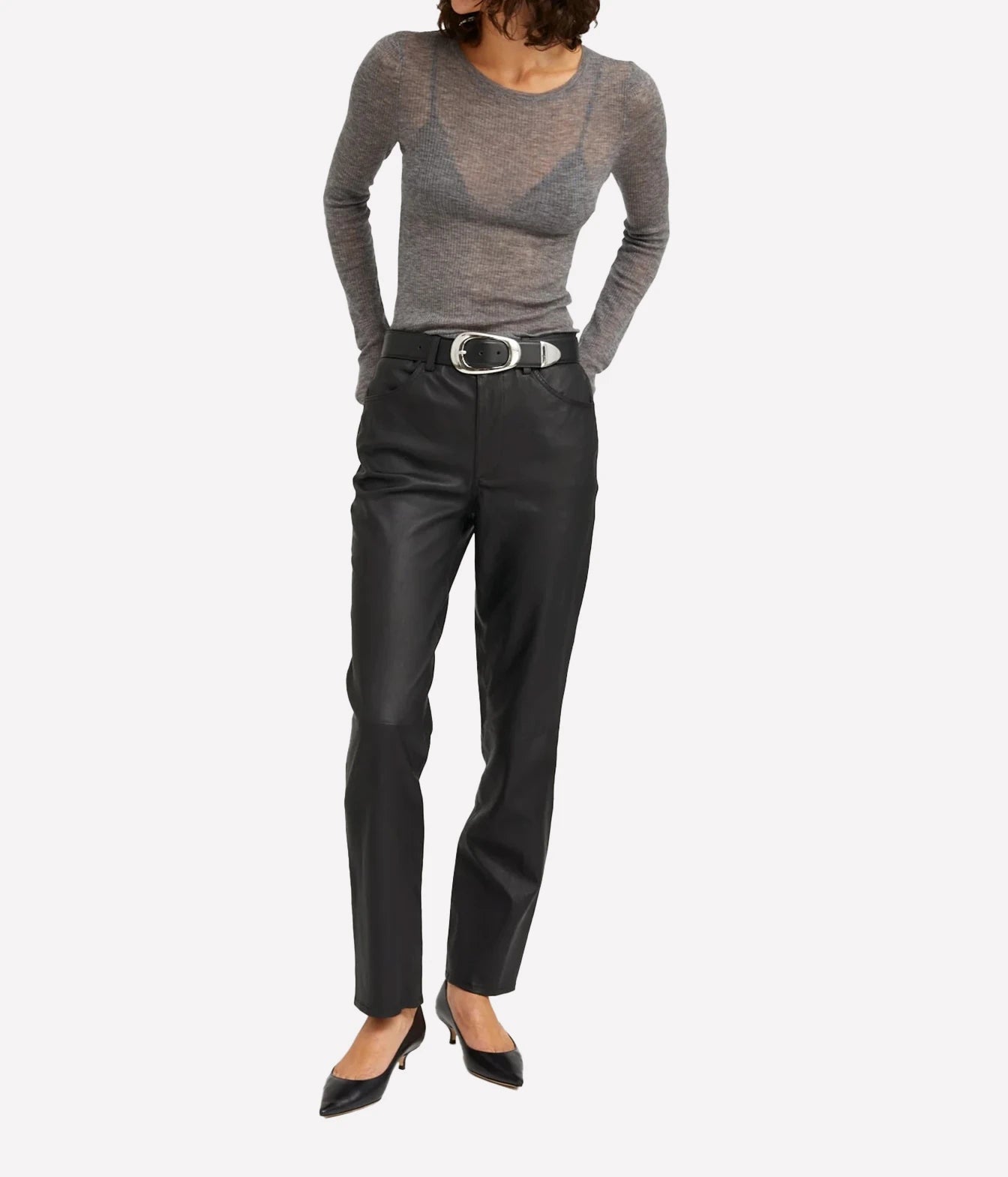 A model wearing a gray long-sleeve knit top, black leather trousers with a silver-belt, and black pointed-toe heels, shown against a plain light gray studio background.