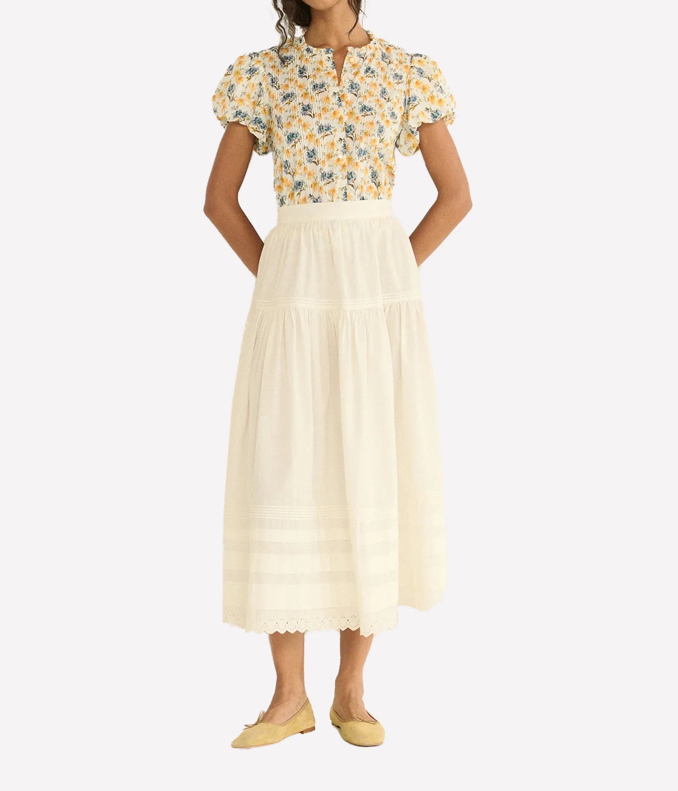 A model wearing a two-piece outfit consisting of a yellow and blue floral short-sleeve blouse and a cream tiered midi skirt, paired with yellow flats, against a light gray background.