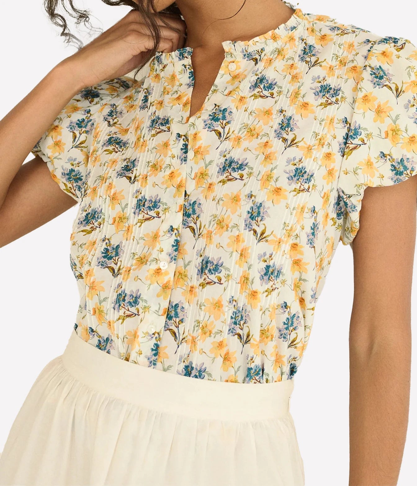 A close-up of a woman wearing a short-sleeve blouse with a yellow and blue floral print on a cream background, paired with a cream skirt.