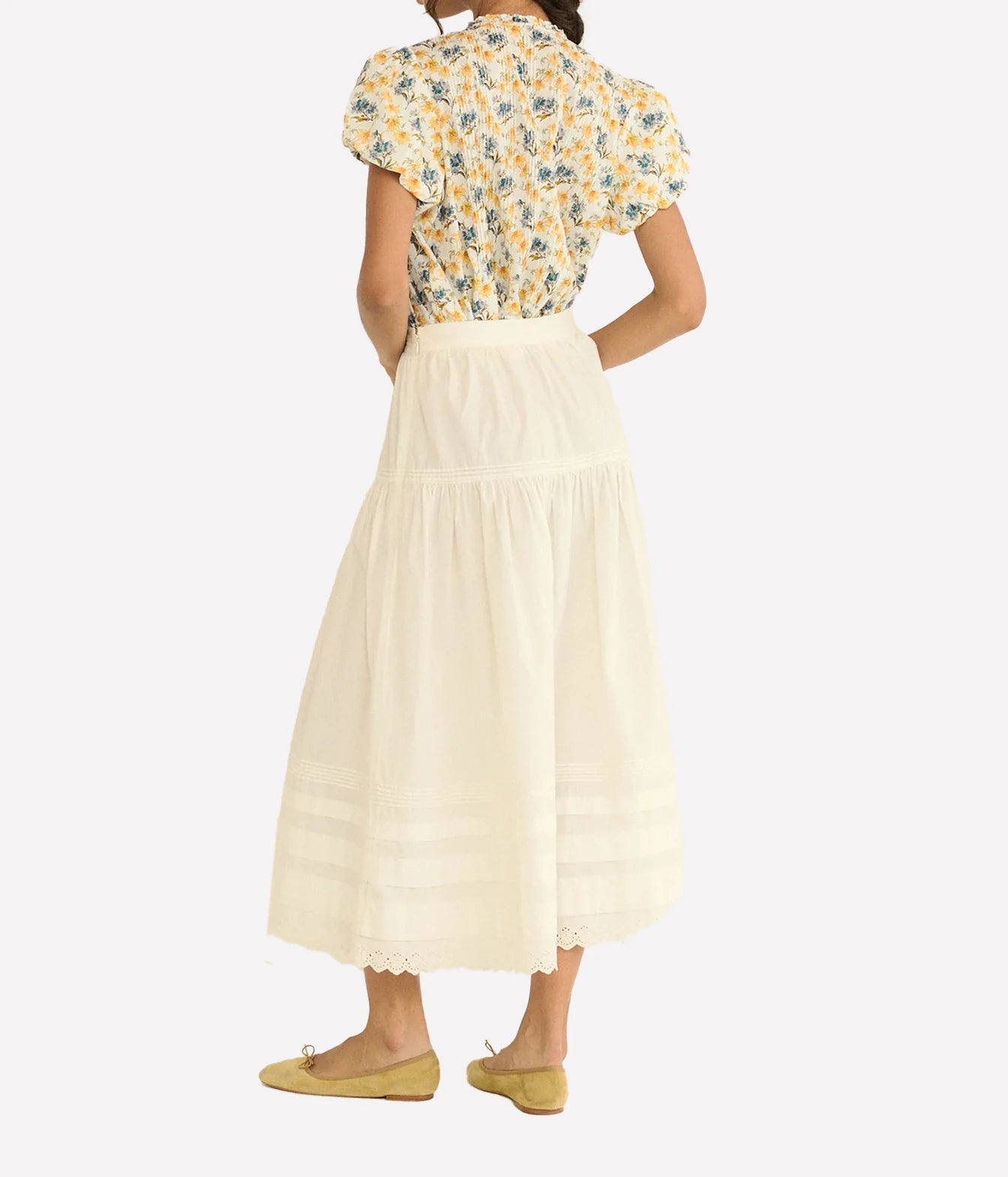 A woman shown from the back wearing a cream midi skirt with horizontal embroidered stripes and a yellow floral short-sleeve blouse, paired with yellow flats, against a light gray background.