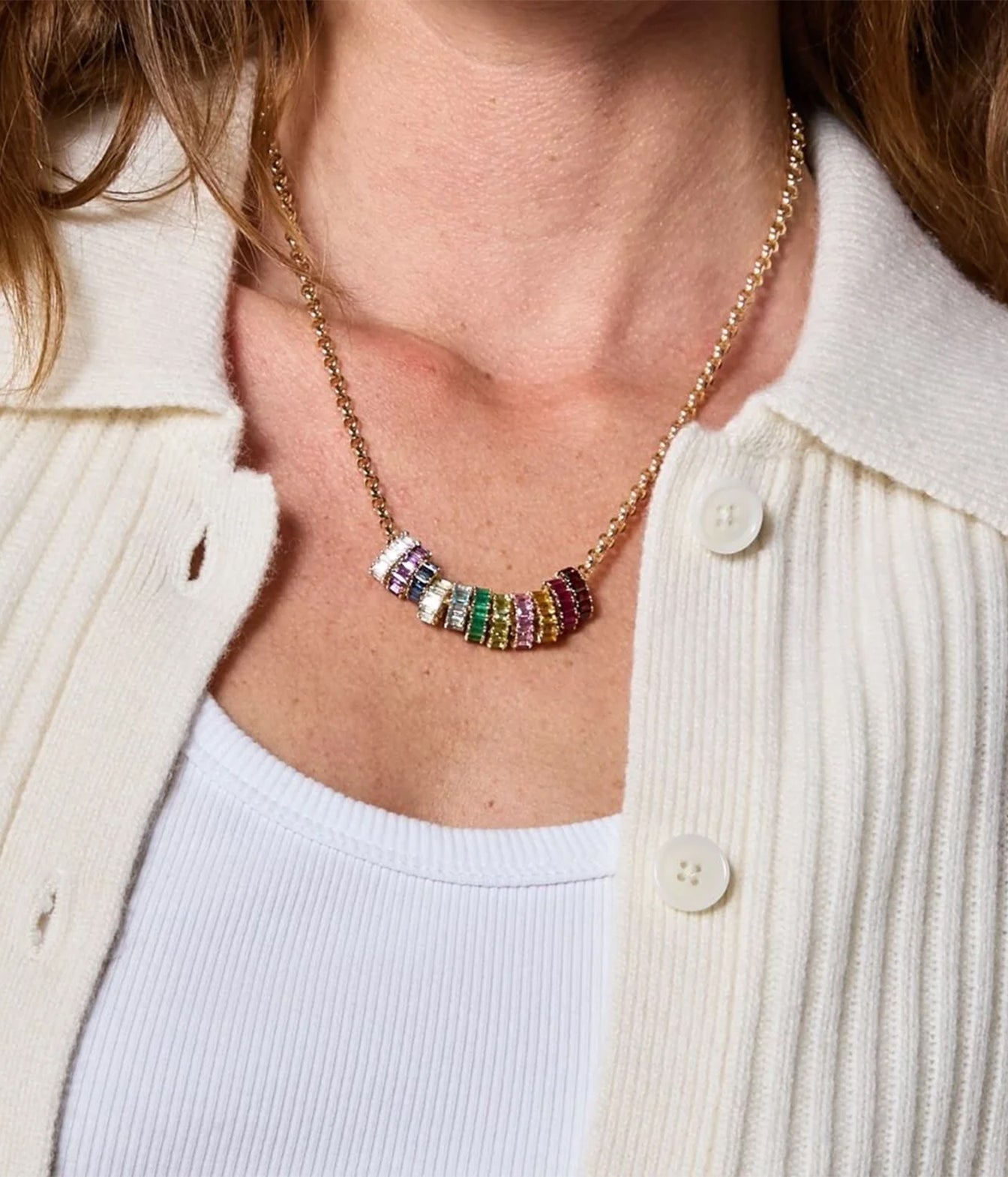 A close-up of a gold chain necklace with a cluster of colorful cylindrical beads, worn by a model in a cream ribbed cardigan over a white top.