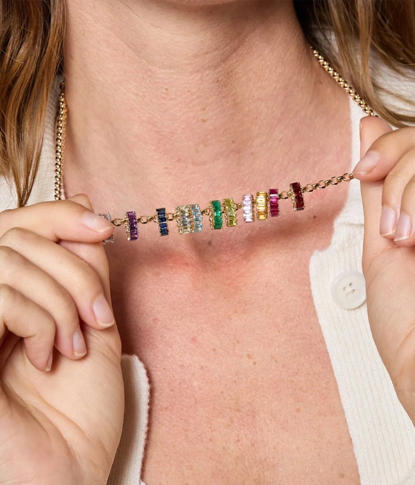 A close-up of a model wearing a delicate gold chain necklace with multiple small, colorful gemstone beads, held in place by her hands against her chest.