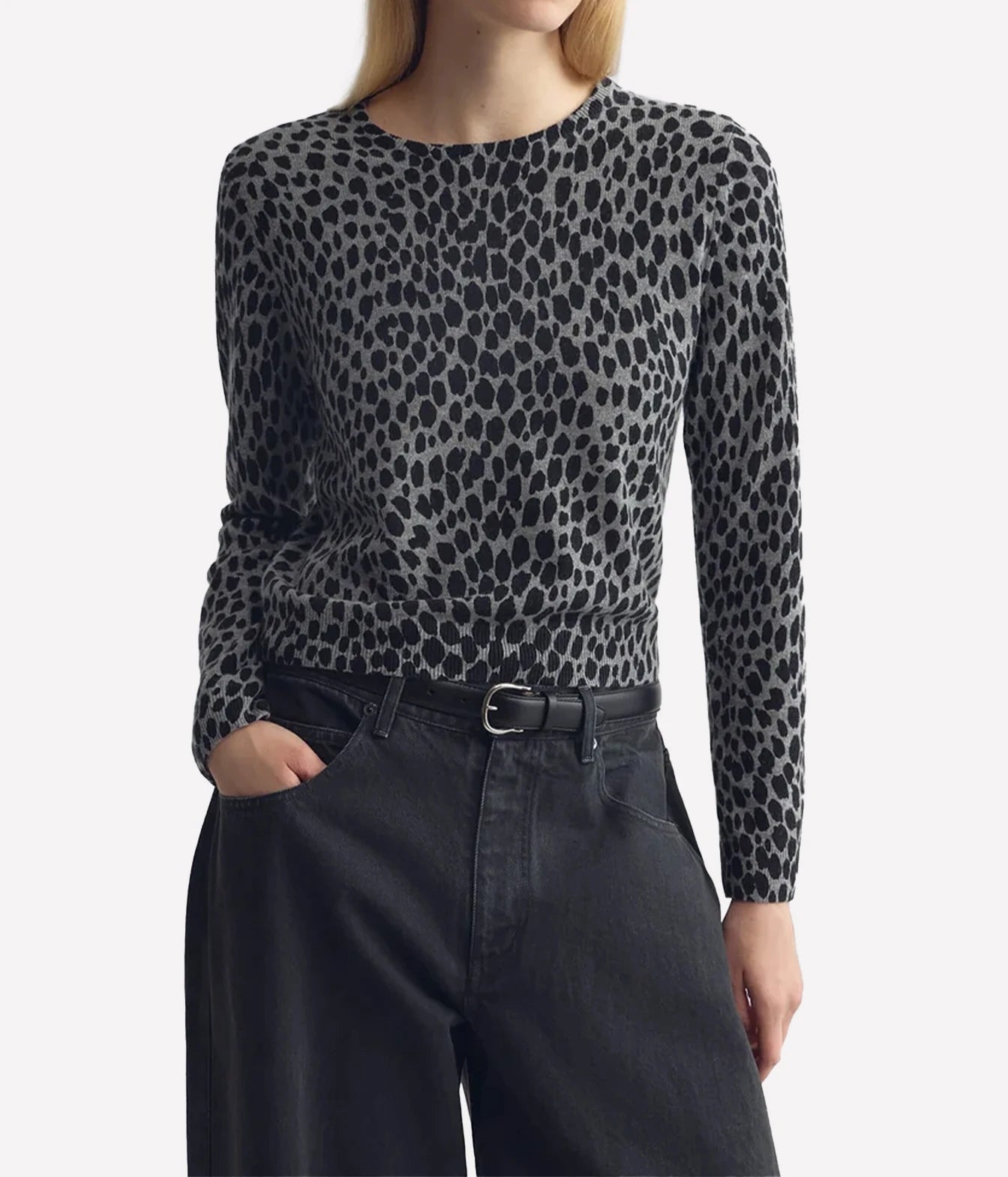 A woman wearing a long-sleeve crewneck knit top with a black and gray leopard print, paired with dark gray jeans and a black belt, against a plain light gray background.