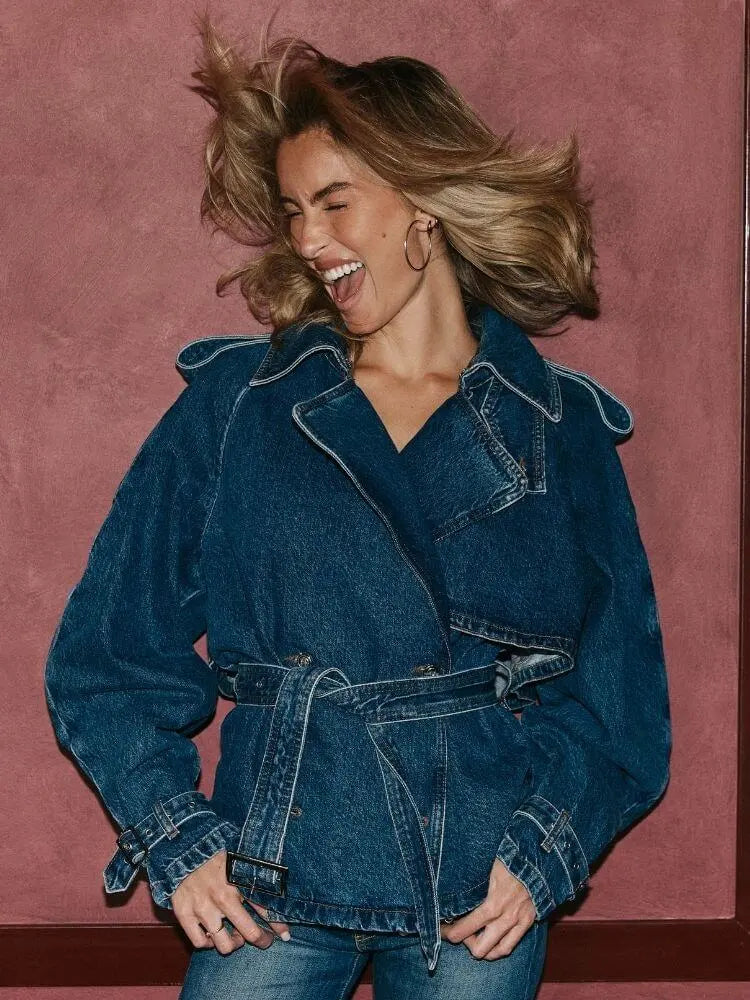 A woman laughing while wearing a dark blue denim belted jacket with contrast stitching, posed against a textured dusty rose wall.