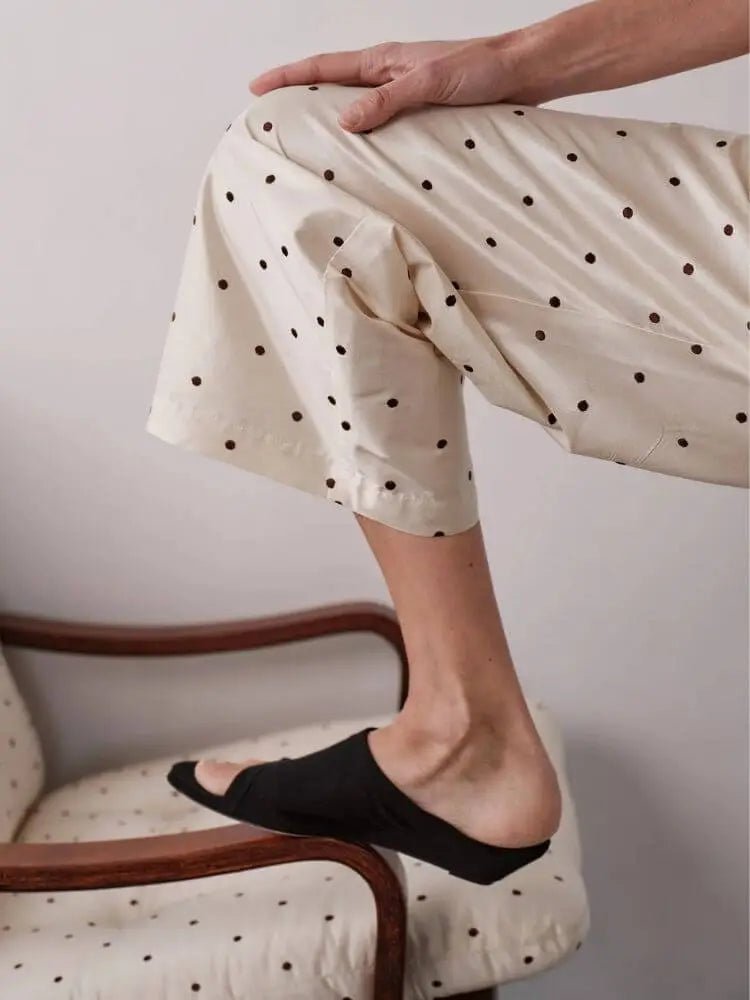 A person seated on a chair wearing loose cream pants with small black polka dots and black slip-on shoes, shown from the waist down in a real indoor setting.