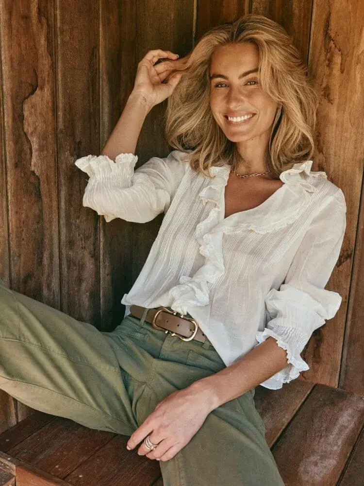 A woman sitting on a rustic wooden bench wearing a white ruffled long-sleeve blouse and olive green pants, posing against a dark wood plank wall.