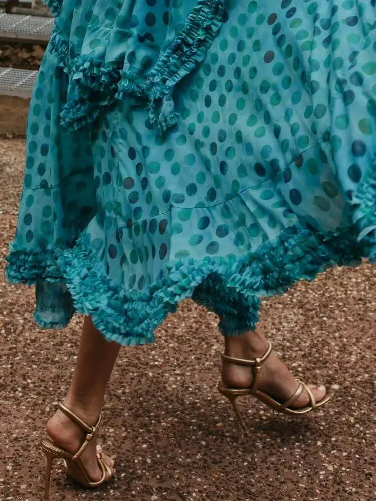 A close-up of a person wearing a flowing teal blue polka dot dress with ruffled trim and gold strappy high-heeled sandals, walking on a gravel path outdoors.