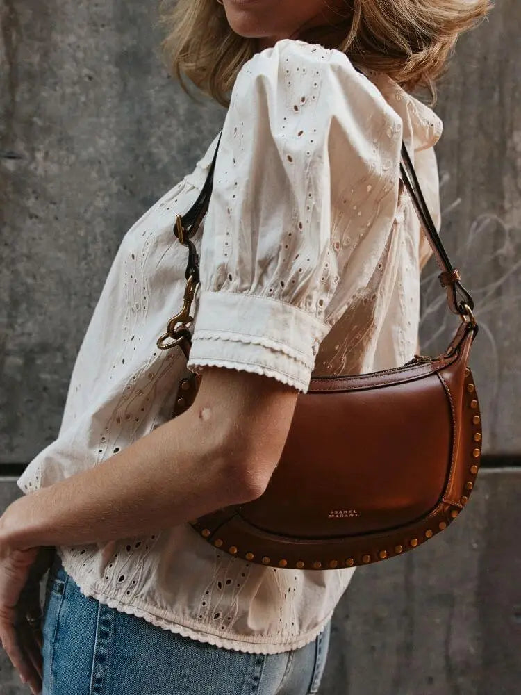 A woman wearing a cream eyelet blouse and blue jeans carries a brown leather crescent-shaped shoulder bag with gold-tone studs and chain detail against a textured gray wall.