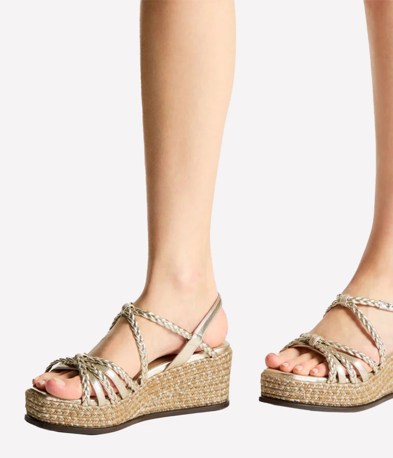 A pair of metallic gold strappy wedge sandals with woven jute platform soles, shown on a model's feet against a light gray background.