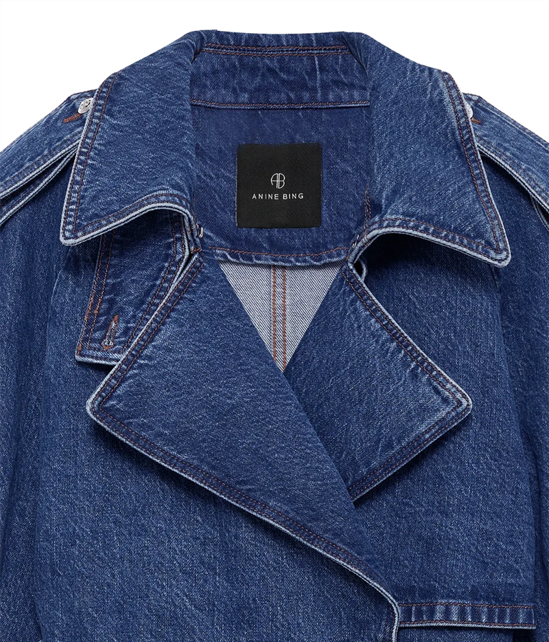 Close-up of a blue denim jacket with a black Anine Bing brand label on the collar, showing the textured denim fabric and contrast stitching.