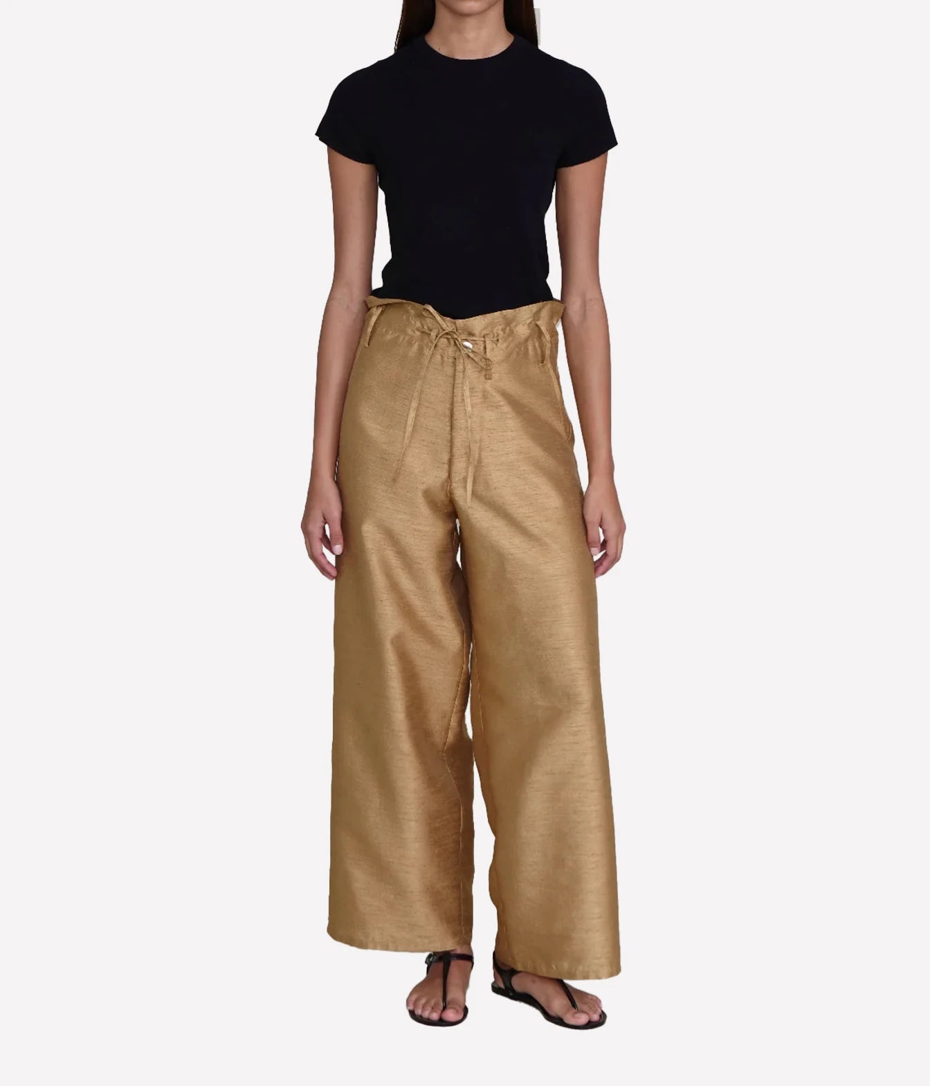 A model wearing a black short-sleeve crewneck top and high-waisted, wide-leg metallic gold trousers with a drawstring waist, paired with black sandals, against a light gray background.
