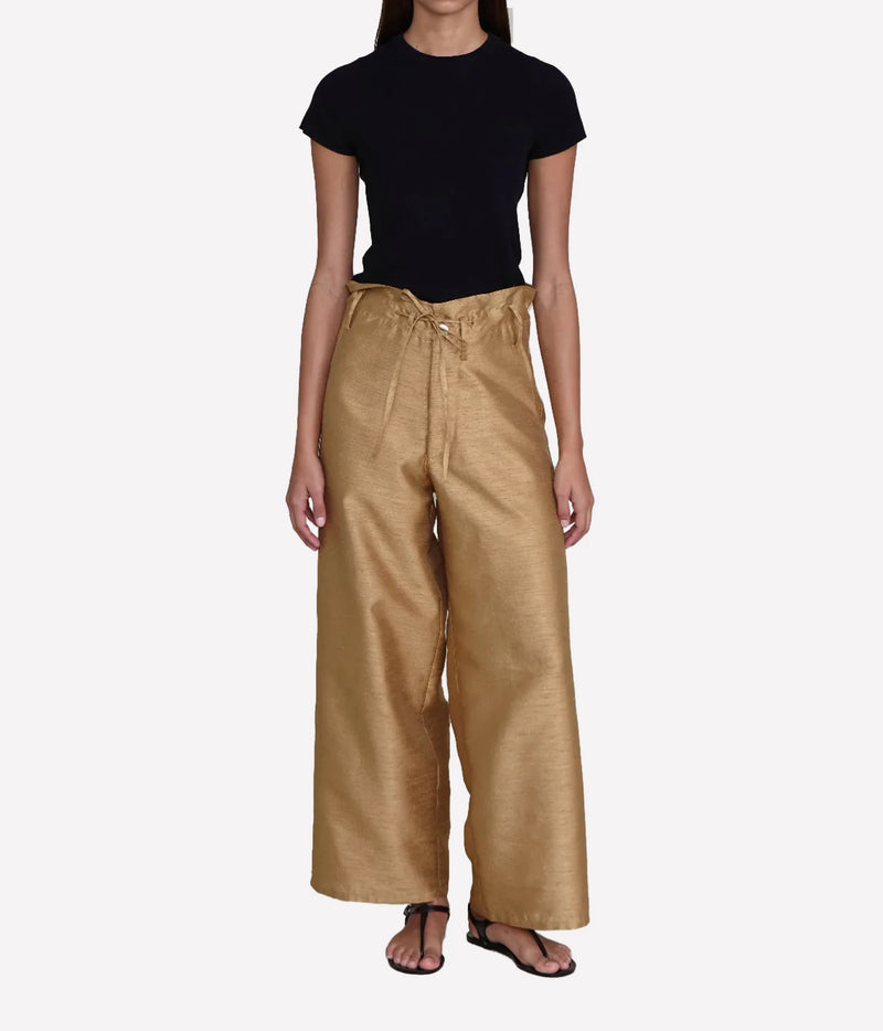 A model wearing a black short-sleeve crewneck top and high-waisted, wide-leg metallic gold trousers with a drawstring waist, paired with black sandals, against a light gray background.