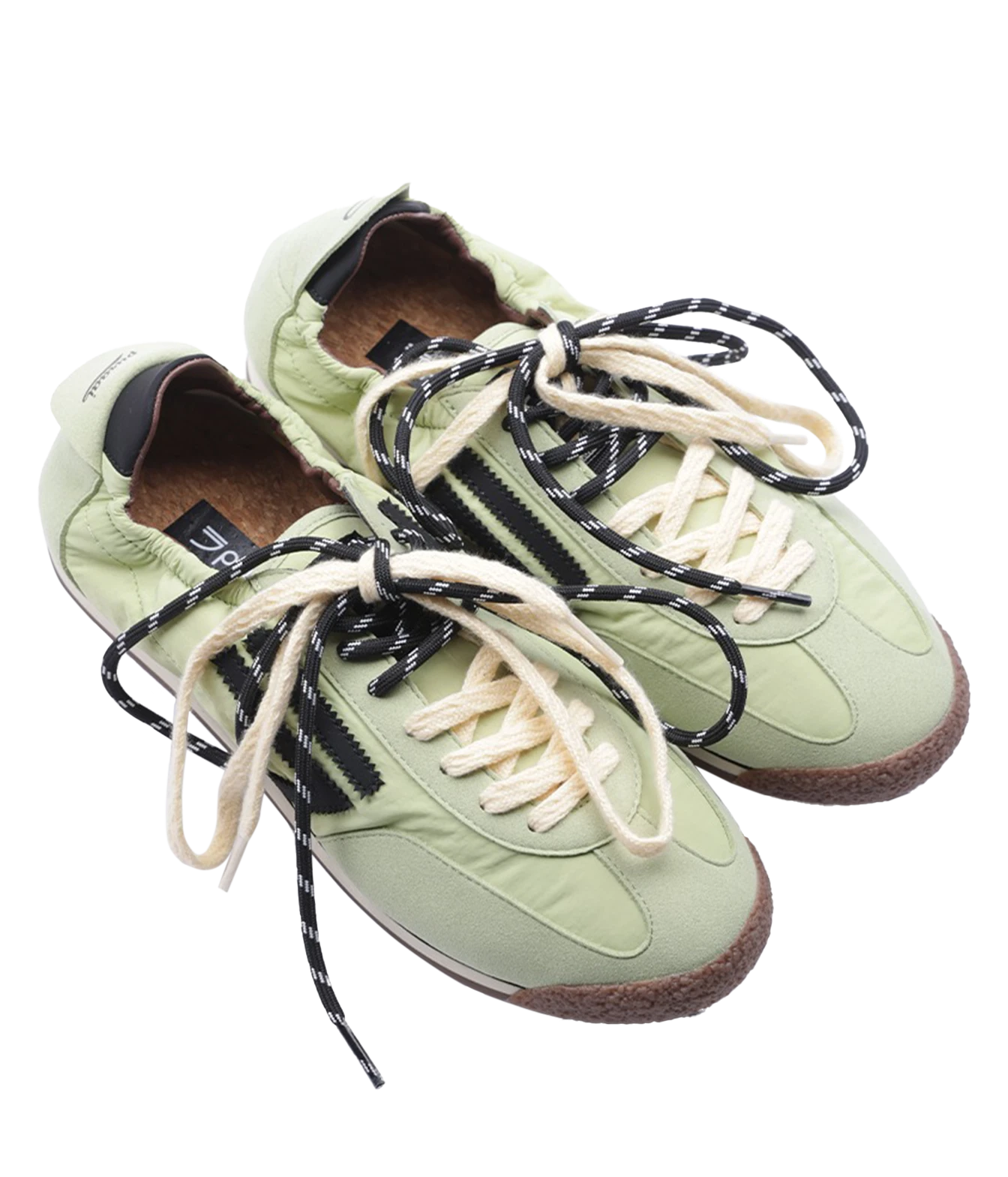 A pair of light sage green sneakers with black and white patterned laces, black side stripes, and brown midsoles, shown on a plain white background.