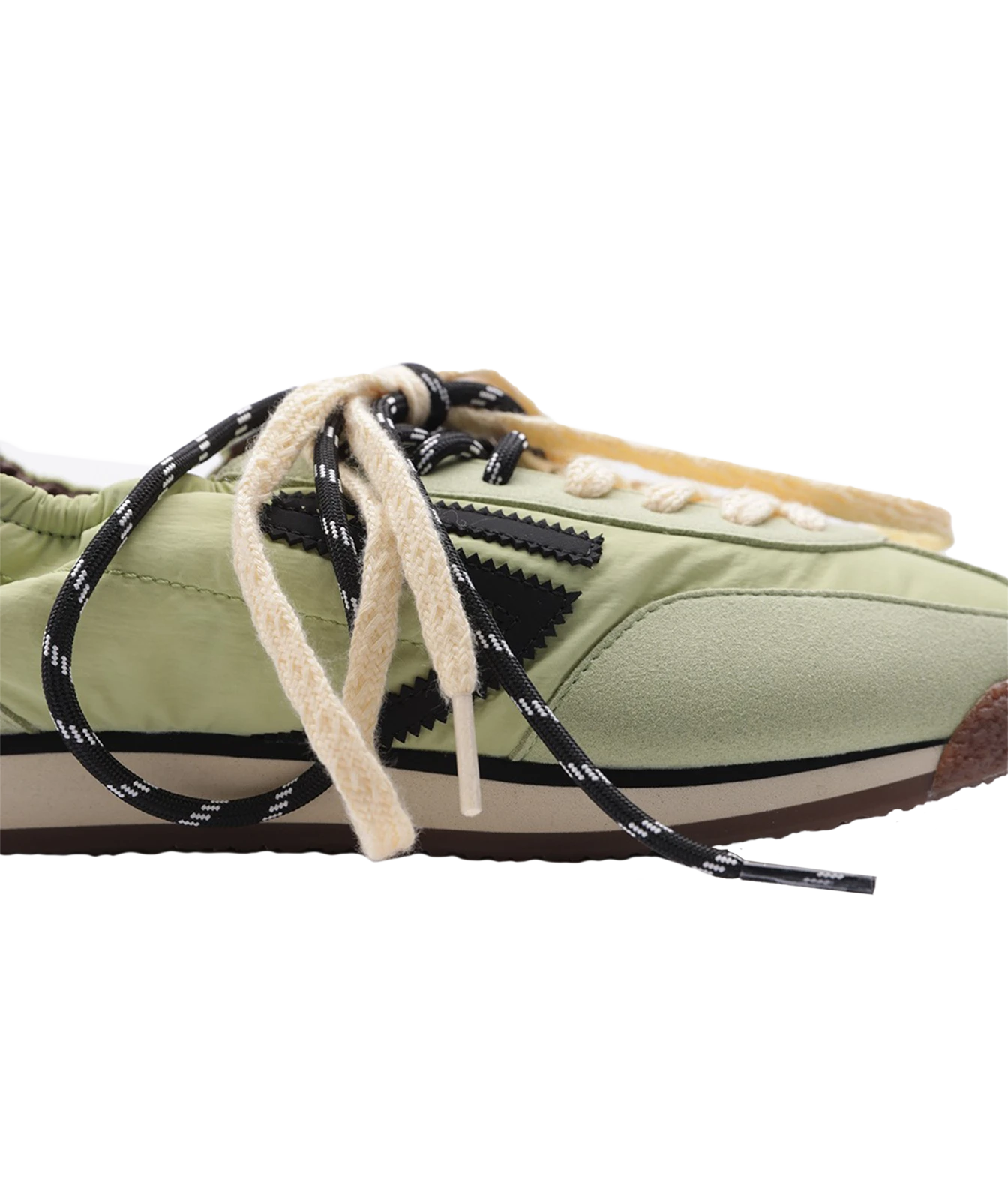 A close-up side view of a low-top sneaker in light olive green, featuring black and white patterned laces, a beige suede toe cap, and a brown gum sole, set against a plain white background.