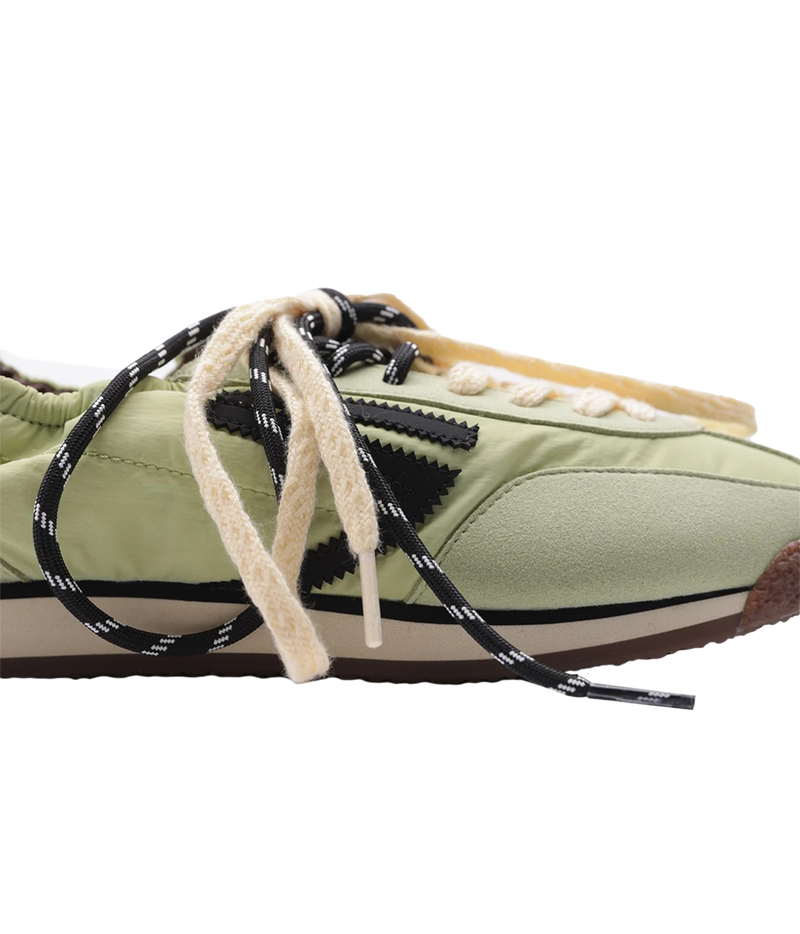 A close-up side view of a low-top sneaker in light olive green, featuring black and white patterned laces, a beige suede toe cap, and a brown gum sole, set against a plain white background.