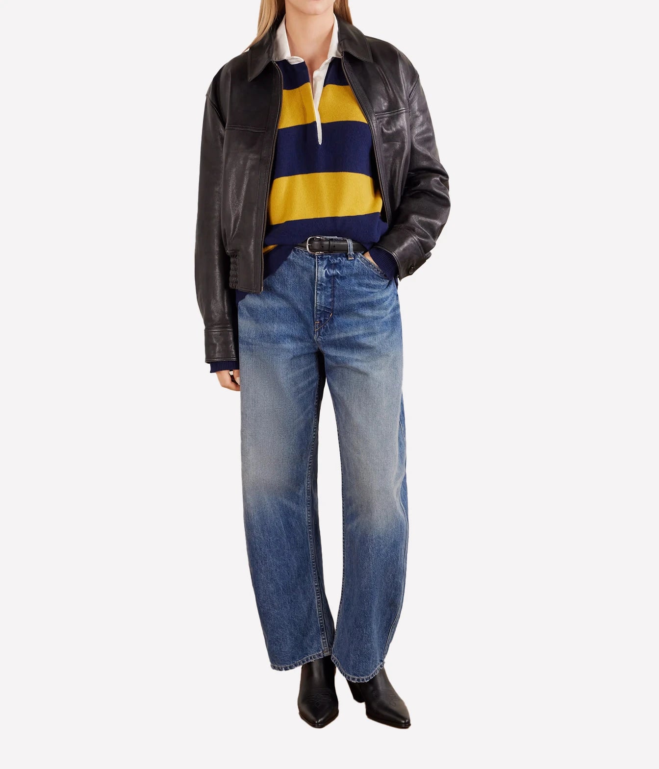 A model wearing a black leather jacket, a navy and yellow striped knit polo shirt, wide-leg faded blue jeans, and black ankle boots, shown against a plain light gray studio background.