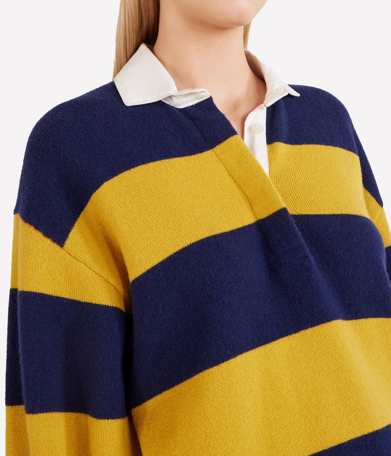 Close-up of a person wearing a knit rugby-style sweater with wide navy and mustard yellow horizontal stripes over a white collared shirt, against a light gray background.