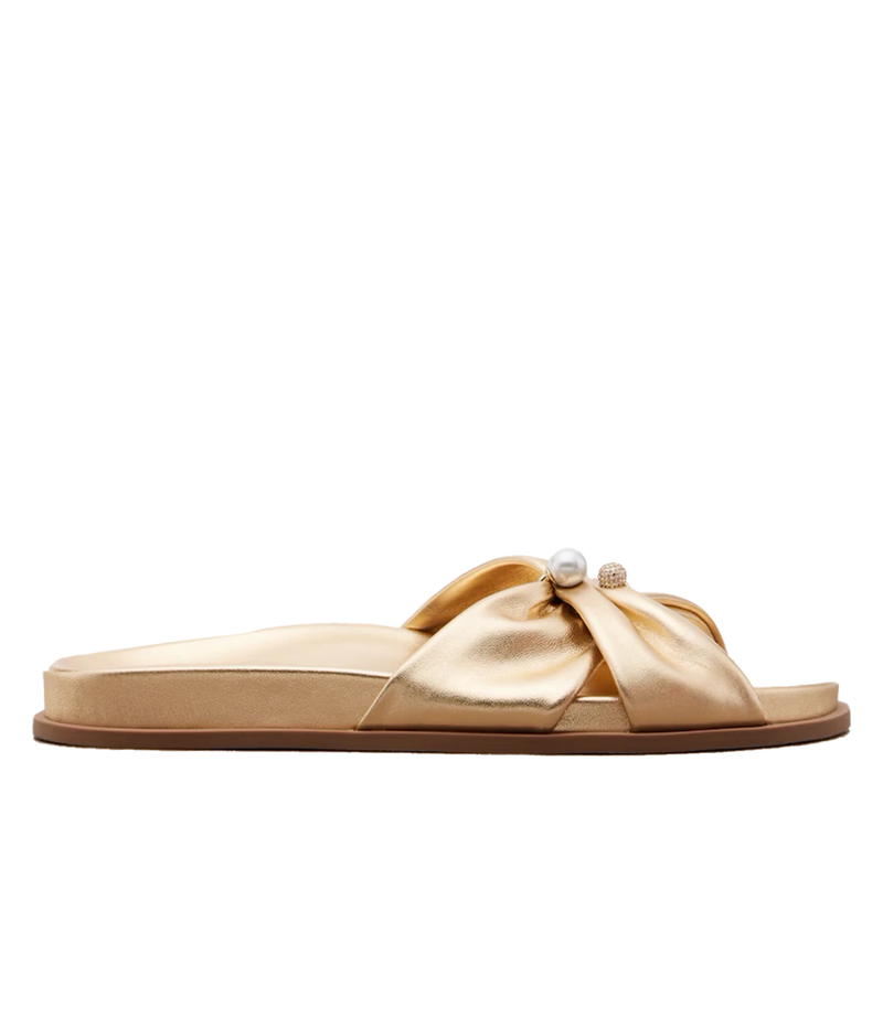 A single flat slide sandal in metallic gold satin, featuring a wide criss-cross bow design across the top adorned with a small white pearl and a tiny gold bead, shown against a plain white background.