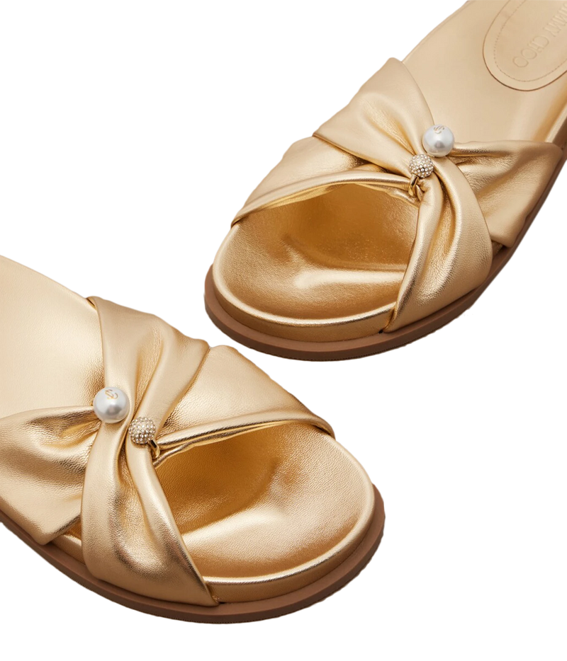 A pair of metallic gold slide sandals with satin bow details and a single white faux-pearl embellishment on the front, shown on a clean white background.