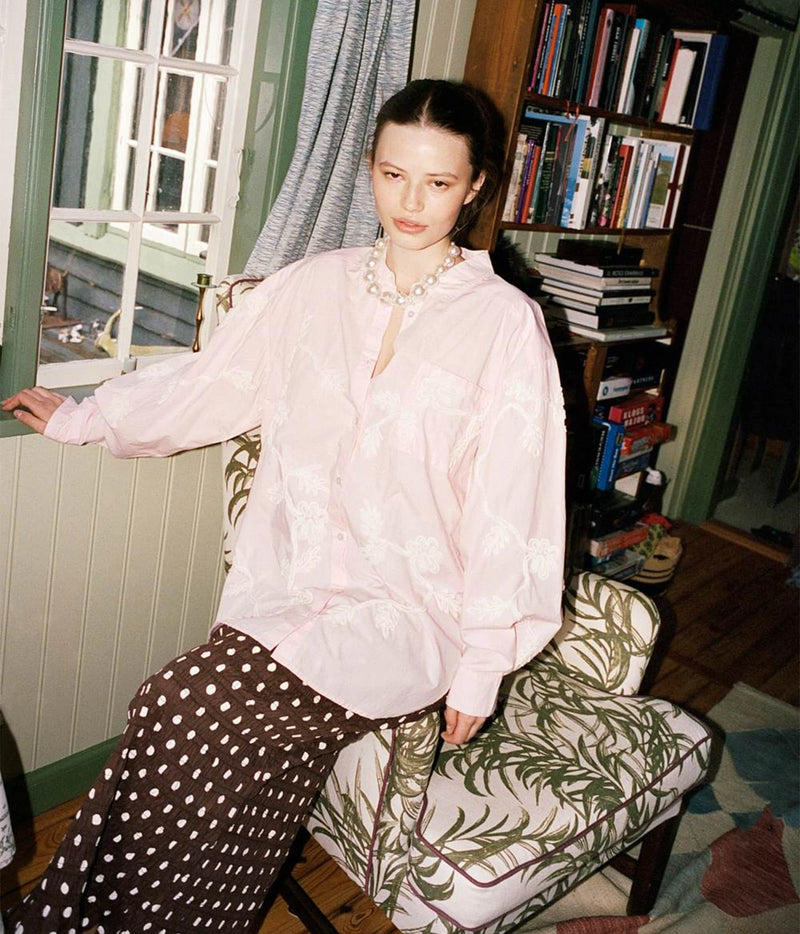 A woman sits in a patterned armchair in a cozy, book-filled room, wearing a light pink embroidered long-sleeve blouse and brown polka-dot trousers.
