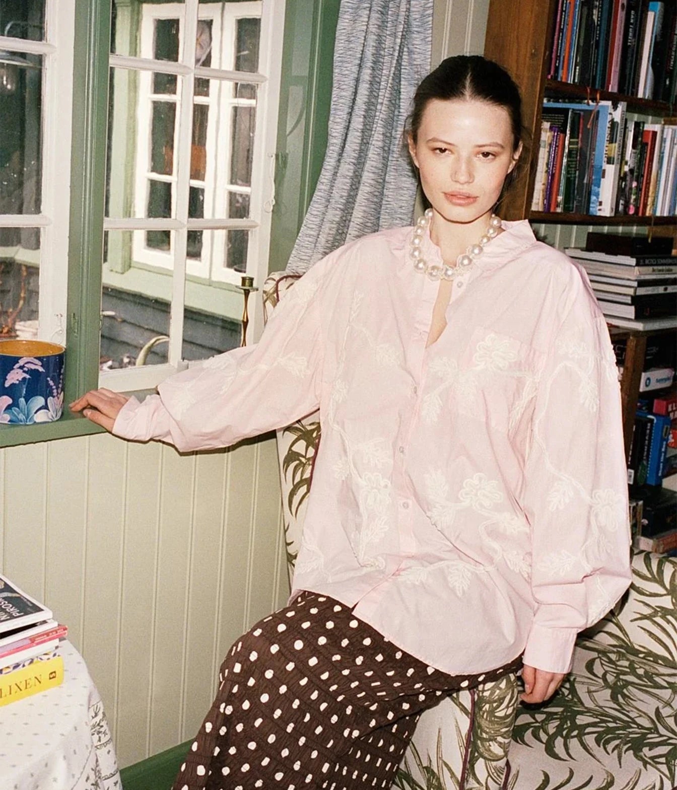 A woman seated indoors wearing a light pink embroidered blouse and dark brown polka-dot trousers, with a pearl necklace, in front of a window and bookshelves.